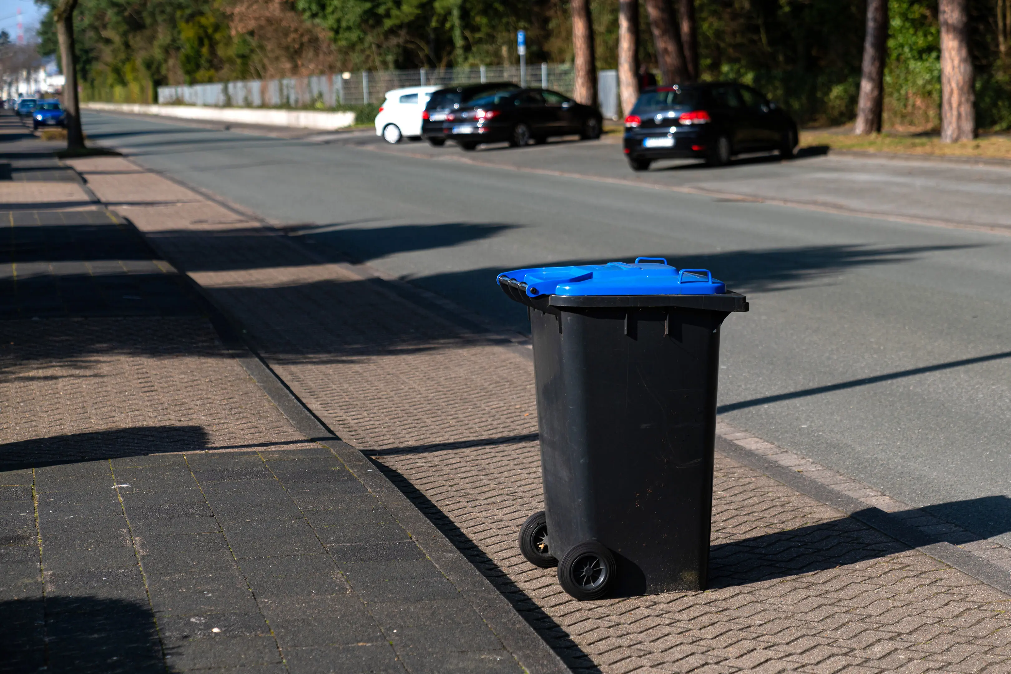 A black trash can with a blue lid on the edge of the street. Parked cars in the background.