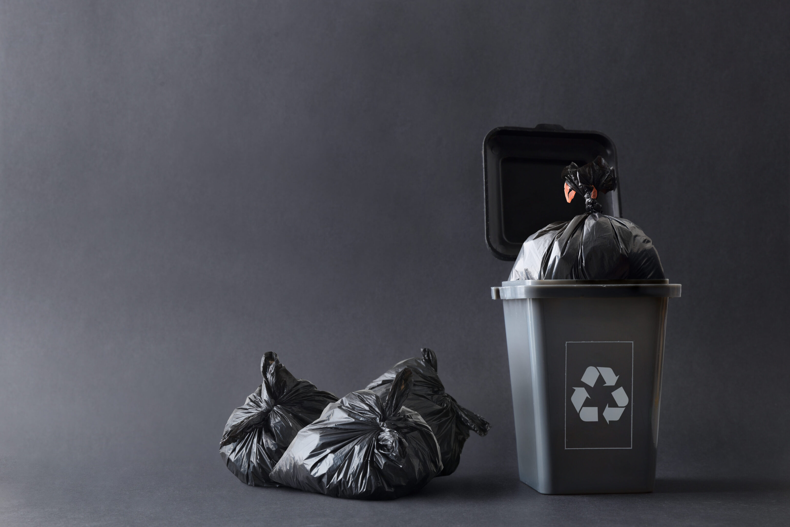 Gray generic garbage recycling container with bags isolated gray Gray trash container with recycling symbol filled with garbage bag and other bags next to it on the ground isolated with gray gradient background. Front view.